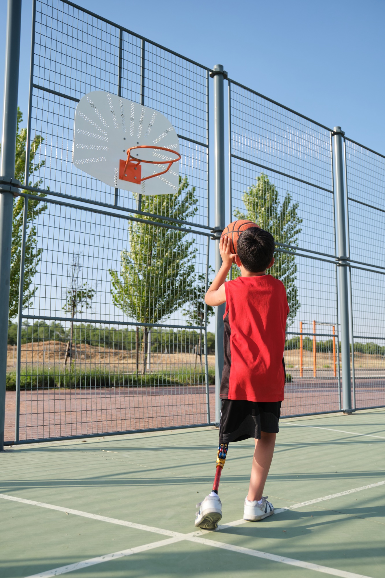 Back view of a basketball kid player with a leg prosthesis throwing the ball to the basket